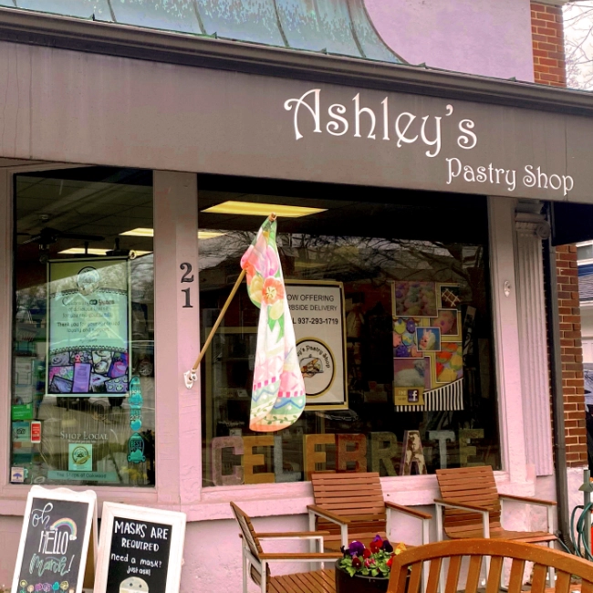 Front view of Ashley's Pastry Shop, a charming local bakery, with outdoor seating and various colorful decorations and posters in the windows.