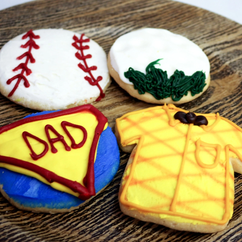 Four decorated cookies on a wooden surface: a baseball, a white round cookie with green icing, a DAD superhero badge, and a yellow shirt with an orange grid pattern.