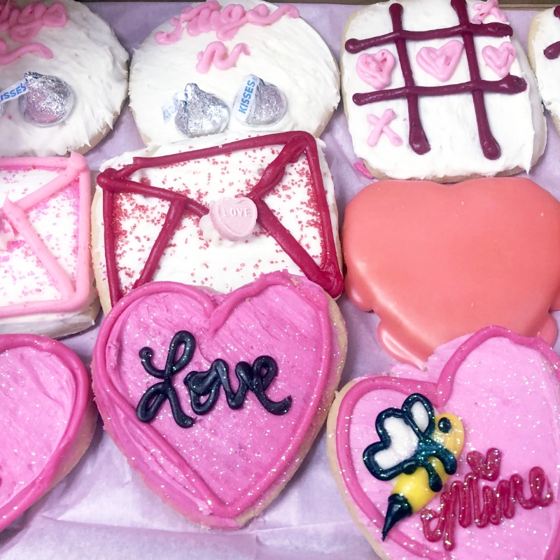 A pink and white assortment of Valentine's Day-themed cookies, including hearts, love letters, and a tic-tac-toe design with candy decorations.