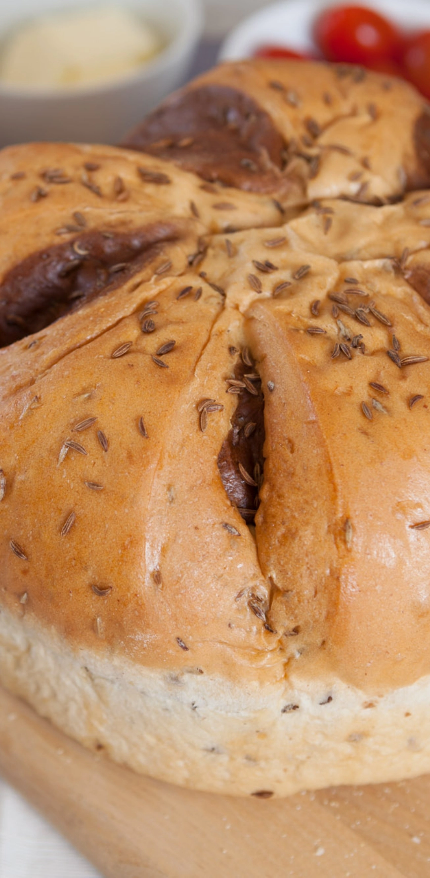 Close-up of a round loaf of bread with a golden-brown crust, sprinkled with caraway seeds, showing deep scored sections. Background slightly blurred with a hint of cherry tomatoes and a small dish.