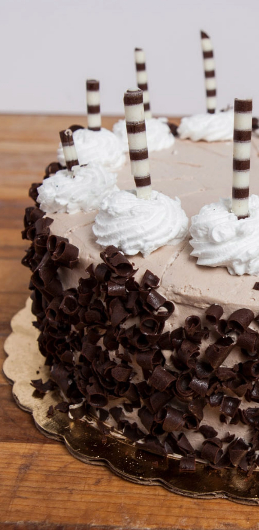 A chocolate cake topped with five whipped cream swirls and striped stick decorations, garnished with chocolate shavings around the sides, placed on a wooden surface.