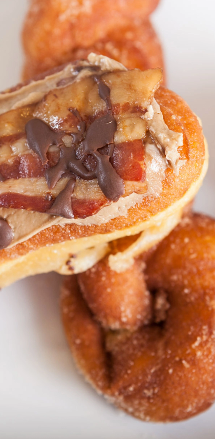 A close-up of a donut topped with bacon, chocolate drizzle, and what appears to be peanut butter. Other plain sugar-coated donuts can be seen in the background.