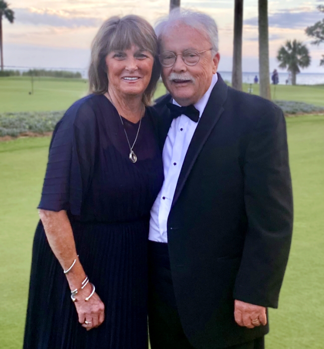 An older couple dressed in formal attire stands together, smiling, with a scenic background of grassy lawn, palm trees, and a sunset sky.
