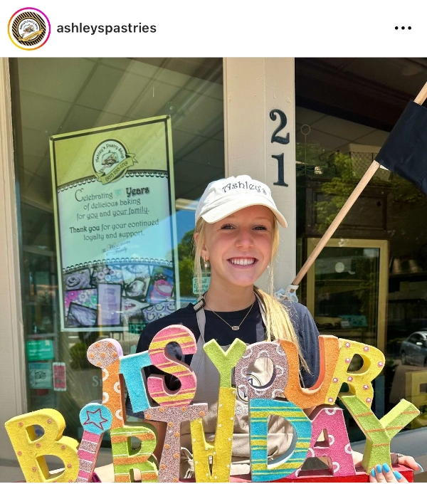 A young woman wearing an "Ashley's" cap and apron holds colorful, sparkly letters spelling "IS YOUR BIRTHDAY" in front of the local bakery, with the number 21 on its door.