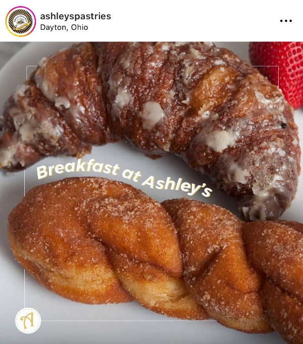 A glazed croissant and a piece of cinnamon-twisted pastry, made fresh at the local bakery, sit on a white plate with a strawberry in the background. Text overlay reads "Breakfast at Ashley's.