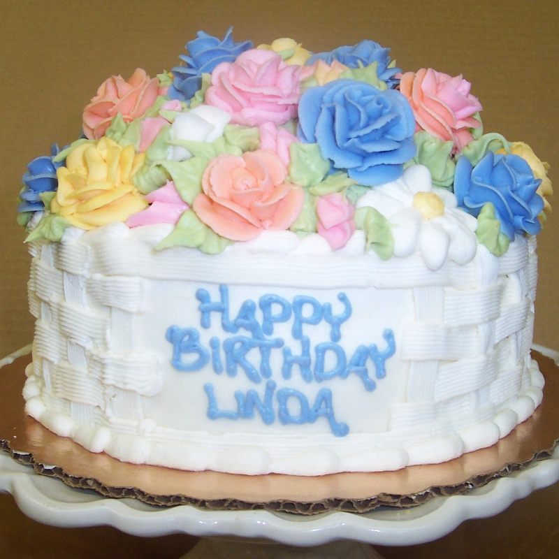 A white frosted cake with a woven basket design, decorated with colorful icing flowers on top. The cake has "Happy Birthday Linda" written in blue icing.
