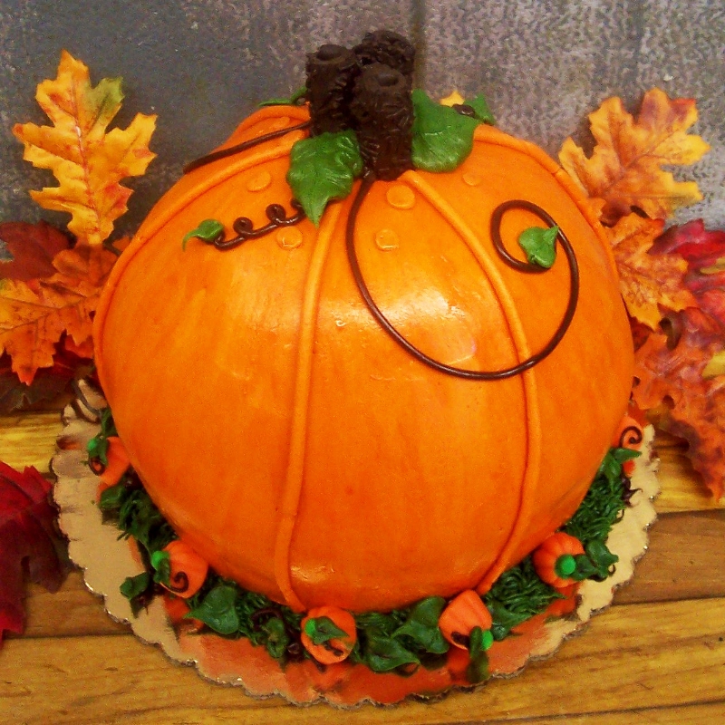 Round cake designed to resemble a pumpkin, with orange frosting, green leaves, and chocolate vine decorations, set against a backdrop of autumn leaves.