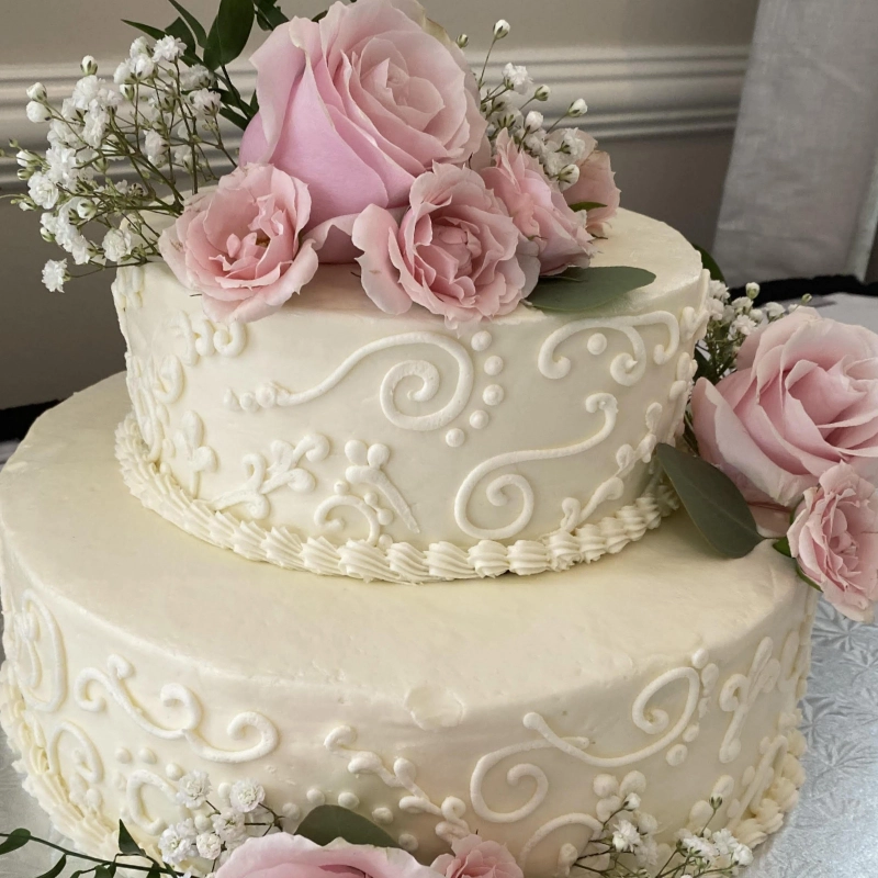 Two-tier white frosted cake decorated with pink roses and white baby's breath flowers, placed on a white tablecloth.
