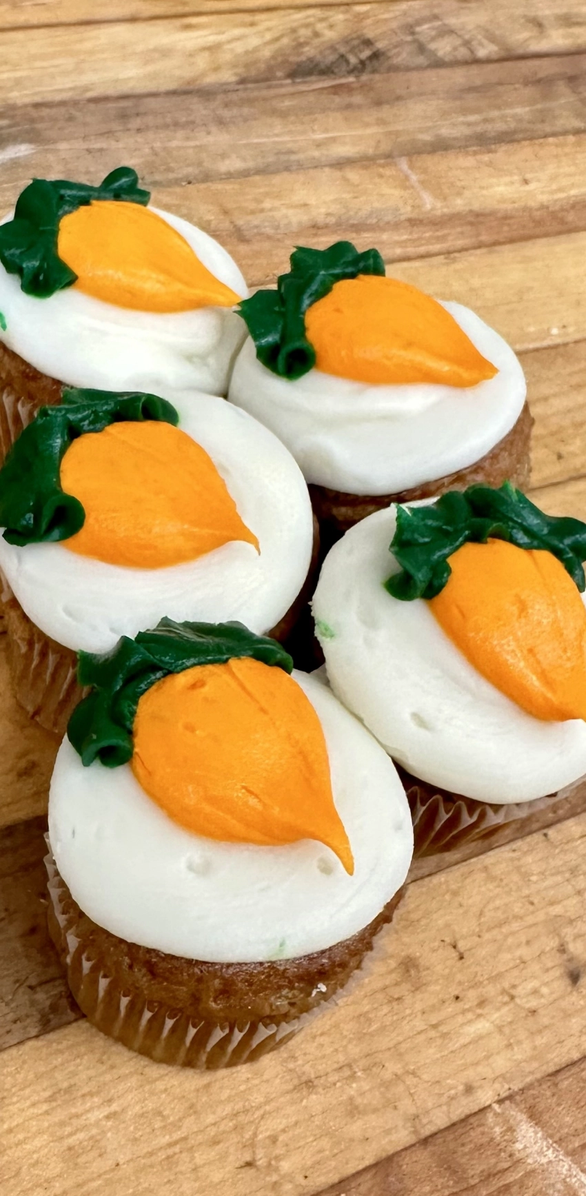 Five cupcakes with white frosting and orange carrot decorations on top, arranged on a wooden surface.