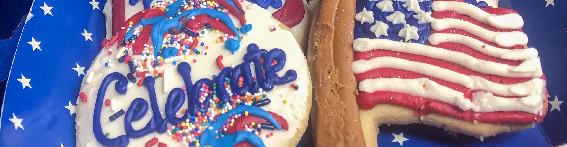Two decorated cookies on a starry blue background: one with "Celebrate" written in icing and colorful sprinkles, the other shaped like a heart with the American flag design, perfect for celebrating the 4th of July.
